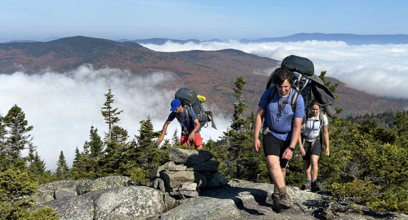 Three people wearing backpacks move over rocky terrain high above the clouds below